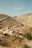 A rugged landscape with rocky hills and sparse vegetation, featuring a dry, arid environment. Three people are seen exploring the area, with one of them accompanied by a dog, adding a sense of adventure to the scene.