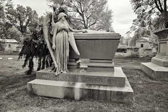 A cemetery scene featuring a large stone tomb with an angelic statue resting against it. The tomb is inscribed with the name of an individual and dates. Surrounding the tomb are trees and other grave markers, creating a solemn and historical atmosphere.