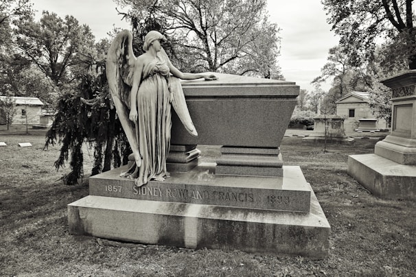 A cemetery scene featuring a large stone tomb with an angelic statue resting against it. The tomb is inscribed with the name of an individual and dates. Surrounding the tomb are trees and other grave markers, creating a solemn and historical atmosphere.