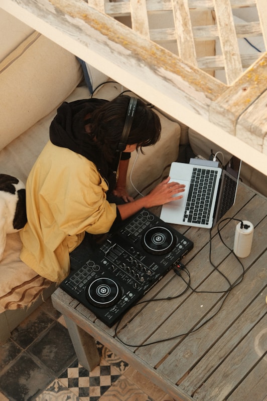 A person wearing headphones is engaged with a laptop while using a DJ controller on a wooden table. The setting appears cozy and relaxed, with a mix of neutral tones. The person is seated on a beige couch, and there's a patterned tile floor visible.