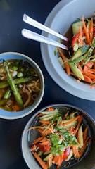 A table setting featuring a variety of vibrant salads and a soup. The salads are composed of fresh vegetables like carrots, cucumbers, tomatoes, and greens, garnished with sesame seeds. The soup bowl contains broth, green vegetables, and asparagus. The dishes are arranged aesthetically in ceramic bowls with metal cutlery beside them.