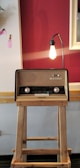 A vintage radio placed on a wooden stand is illuminated by a bare bulb lamp extending from its back. The background features a two-tone wall with a deep red and cream color scheme, adorned with a few decorative elements, including a small piece of pink fabric.