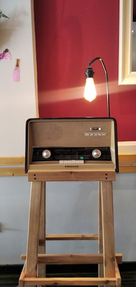 A vintage radio placed on a wooden stand is illuminated by a bare bulb lamp extending from its back. The background features a two-tone wall with a deep red and cream color scheme, adorned with a few decorative elements, including a small piece of pink fabric.