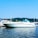 A sleek white yacht is docked in a marina, surrounded by several other boats. The water is calm, and there are lush green hills in the background under a clear blue sky.