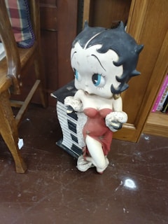 A cartoon-like sculpture of a character with large eyes, wearing a red dress and positioned next to a piano-shaped object. The setup is indoors on a brown floor, surrounded by wooden furniture, including a shelf filled with books.