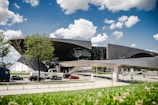 A modern architectural building featuring curved and angular designs, set against a clear blue sky with scattered white clouds. Trees and a well-maintained lawn are in the foreground, while vehicles are visible on the road in the midground, suggesting an urban setting.
