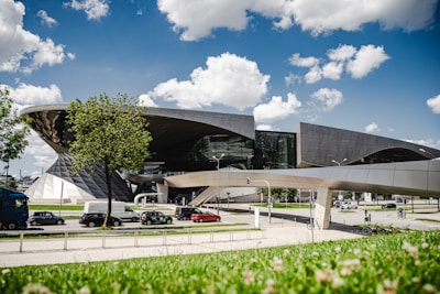 A modern architectural building featuring curved and angular designs, set against a clear blue sky with scattered white clouds. Trees and a well-maintained lawn are in the foreground, while vehicles are visible on the road in the midground, suggesting an urban setting.