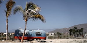 A vintage trailer with a red base and blue upper part is parked on a sandy ground. Two tall palm trees stand nearby against a backdrop of mountains and a clear blue sky. The setting appears to be open and spacious, possibly a remote or desert area.