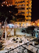 The image captures an archaeological excavation site surrounded by tall buildings at night. The site features stone ruins and remnants of columns, indicating historical architecture. Trees are scattered around the area, and artificial lighting from the buildings creates a warm glow illuminating the scene. The street level is elevated above the excavation, with shops and lights visible.
