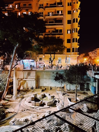 The image captures an archaeological excavation site surrounded by tall buildings at night. The site features stone ruins and remnants of columns, indicating historical architecture. Trees are scattered around the area, and artificial lighting from the buildings creates a warm glow illuminating the scene. The street level is elevated above the excavation, with shops and lights visible.