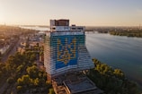 A large, abandoned hotel building features a painted design of a trident in blue and yellow colors. The structure is situated near a river, with an expansive view of the water, cityscape, and bridges in the background. The surrounding area includes industrial sites and lush green trees, basking in the warm light of a setting or rising sun.