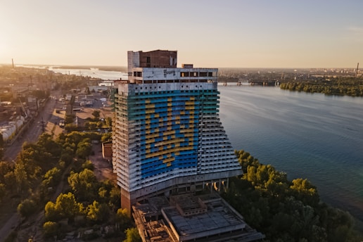 A large, abandoned hotel building features a painted design of a trident in blue and yellow colors. The structure is situated near a river, with an expansive view of the water, cityscape, and bridges in the background. The surrounding area includes industrial sites and lush green trees, basking in the warm light of a setting or rising sun.