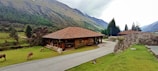 A rustic building with a brown tiled roof situated in a lush, green mountainous landscape. The structure appears to be a visitor center or shop surrounded by grassy fields where a few animals, possibly alpacas or llamas, are grazing. Tall trees and a rocky terrain form the backdrop, with cloudy skies overhead.