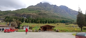 A tranquil mountain landscape with a lodge in the foreground surrounded by lush greenery. A person in a red coat walks on a paved surface, and several parked cars are visible. In the background, a waterfall flows down the mountain with dense forestation on the slopes.