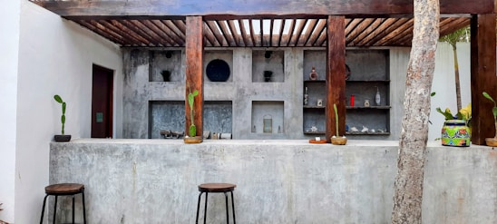 An outdoor bar area with a concrete counter and wooden stool seating. The back wall displays various decorative items, including vases and small plants. The structure is supported by wooden beams, and a colorfully painted planter is placed on the counter. Minimalist design with potted plants adds an earthy and artistic touch.
