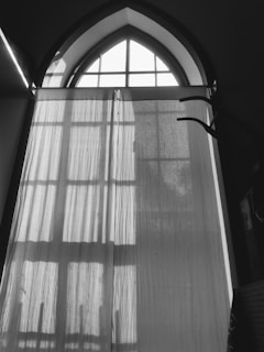 A tall, arched window with grilles is covered by a sheer curtain, allowing filtered light to pass through and cast shadows on the fabric. The scene is framed by the window's architecture and nearby furniture or decor.