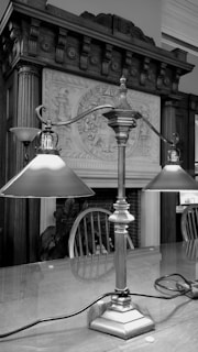 A table lamp with two shades sits on a polished wooden table in a room with ornate wooden paneling and a decorative fireplace. The background features detailed carvings and a classic wooden chair. The scene appears to be part of a traditional interior setting.