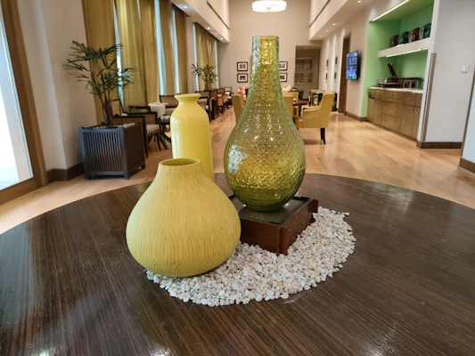The image depicts a modern indoor setting featuring a round wooden table adorned with three decorative vases of varying shapes and sizes. The vases are placed on a bed of small white pebbles. Behind the table, there's a spacious room with wooden flooring, contemporary furnishings, including upholstered chairs and tables. Plants in pots line the perimeter, and large windows allow natural light to illuminate the space. The color palette is warm with a mix of natural and earthy tones.