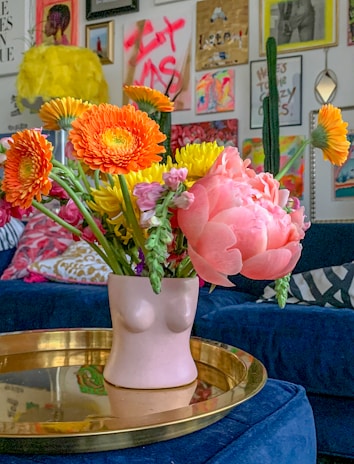 A decorative room with a vase shaped like a human torso on a gold tray, containing vibrant flowers including orange gerberas and pink peonies. The background displays eclectic wall art with colorful paintings and a cactus. The room has a stylish, bohemian vibe with a mix of textures and colors.