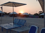 A beachside setting with a calm atmosphere featuring arranged sun loungers under umbrellas. The sun is setting in the background, casting a warm glow over the area. Tents with white tops are positioned on the sand, surrounded by trees.