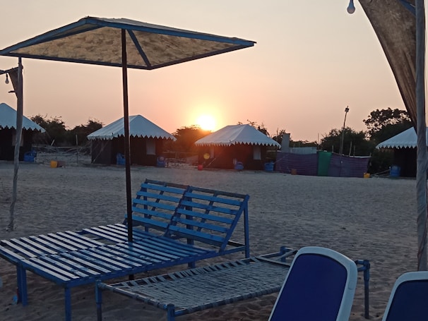A beachside setting with a calm atmosphere featuring arranged sun loungers under umbrellas. The sun is setting in the background, casting a warm glow over the area. Tents with white tops are positioned on the sand, surrounded by trees.