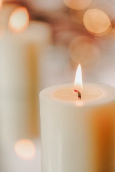 A close-up of a lit candle with a soft, warm glow. The light reflects gently off the off-white wax, while the background features blurred, circular bokeh lights in shades of gold and amber.