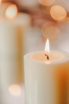 A close-up of a lit candle with a soft, warm glow. The light reflects gently off the off-white wax, while the background features blurred, circular bokeh lights in shades of gold and amber.