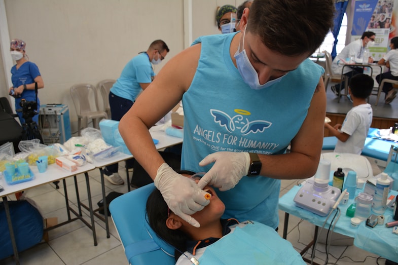 A person in a blue shirt with the logo 'Angels for Humanity' provides dental care to a young patient lying on a dental chair. The setting appears to be a makeshift dental clinic with blue medical equipment and supplies on tables. Other team members in similar attire are visible in the background, some attending to other patients.