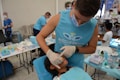 A person in a blue shirt with the logo 'Angels for Humanity' provides dental care to a young patient lying on a dental chair. The setting appears to be a makeshift dental clinic with blue medical equipment and supplies on tables. Other team members in similar attire are visible in the background, some attending to other patients.
