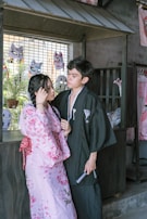 A couple dressed in traditional Japanese clothing stands close together in an area decorated with masks and traditional items. The woman wears a pink floral kimono, and the man wears a dark kimono with subtle patterns. Behind them are colorful Japanese masks and plants, adding cultural ambiance to the setting.