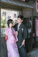 A couple dressed in traditional Japanese clothing stands close together in an area decorated with masks and traditional items. The woman wears a pink floral kimono, and the man wears a dark kimono with subtle patterns. Behind them are colorful Japanese masks and plants, adding cultural ambiance to the setting.