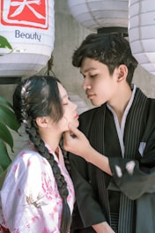 A young couple is dressed in traditional Japanese clothing, standing closely under large paper lanterns. The woman has a braided hairstyle and wears a floral-patterned kimono, while the man in a dark kimono gazes at her, gently touching her chin. Sunlight filters through, creating a soft, intimate atmosphere.
