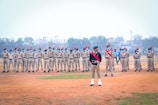 A group of uniformed individuals stands in formation on an outdoor field. They are wearing beige uniforms with green berets adorned with red plumes. Some members have sashes in red, blue, and white, and they all appear to be part of a military or cadet drill. The background is a cloudy sky with some trees and buildings, and a few people are walking in the distance.