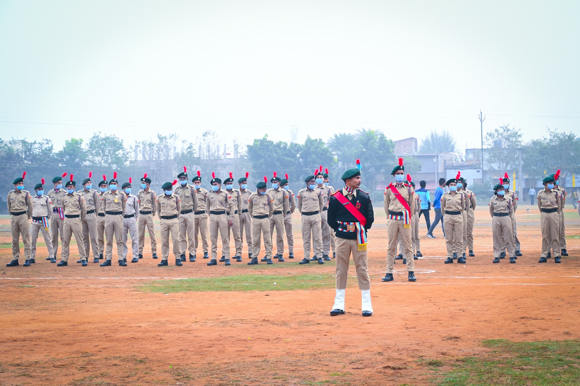 A group of uniformed individuals stands in formation on an outdoor field. They are wearing beige uniforms with green berets adorned with red plumes. Some members have sashes in red, blue, and white, and they all appear to be part of a military or cadet drill. The background is a cloudy sky with some trees and buildings, and a few people are walking in the distance.
