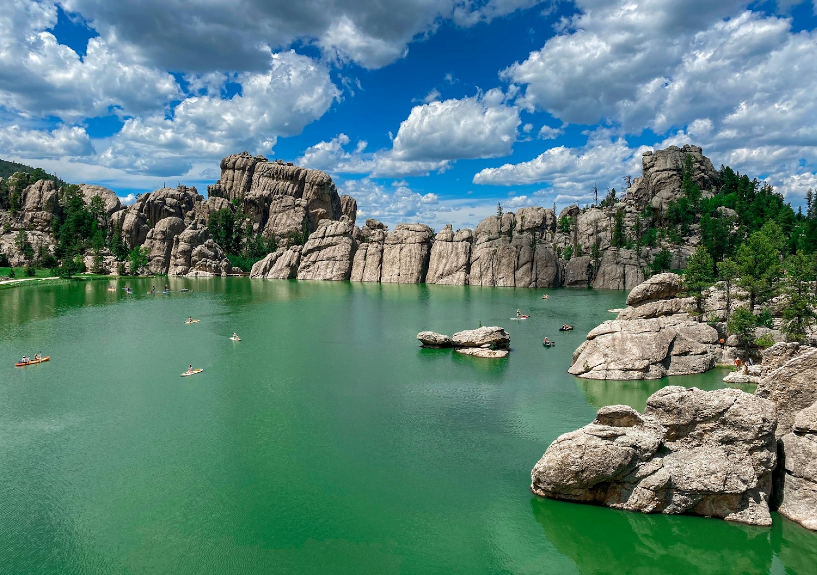 Aerial view of a green lake dotted with kayaks and paddleboards, enclosed by rounded granite cliffs and pine trees under a cloud-filled blue sky