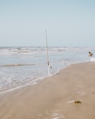 A fishing rod is planted upright in the sand near the edge of the water on a beach. The waves are gently rolling onto the shore. In the background, a person is crouched down near the water, seemingly engaged in an activity.