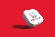 A 3D representation of the Honda logo is centered against a vibrant red background. The logo consists of a silver 'H' emblem above the brand name 'Honda' written in bold red letters, embossed on a white square-shaped surface that casts a shadow, suggesting a floating appearance.
