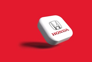 A 3D representation of the Honda logo is centered against a vibrant red background. The logo consists of a silver 'H' emblem above the brand name 'Honda' written in bold red letters, embossed on a white square-shaped surface that casts a shadow, suggesting a floating appearance.