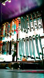 A variety of skateboards are displayed on racks against a window inside a store. The skateboards have various designs and colors, some with colorful graphics and others with solid tones. The lighting creates a vibrant and lively atmosphere with a purple ceiling adding an artistic touch to the space.