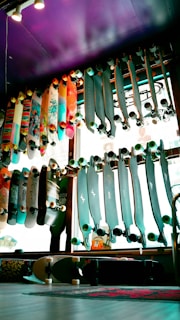 A variety of skateboards are displayed on racks against a window inside a store. The skateboards have various designs and colors, some with colorful graphics and others with solid tones. The lighting creates a vibrant and lively atmosphere with a purple ceiling adding an artistic touch to the space.