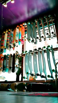 A variety of skateboards are displayed on racks against a window inside a store. The skateboards have various designs and colors, some with colorful graphics and others with solid tones. The lighting creates a vibrant and lively atmosphere with a purple ceiling adding an artistic touch to the space.