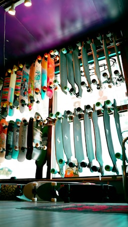A variety of skateboards are displayed on racks against a window inside a store. The skateboards have various designs and colors, some with colorful graphics and others with solid tones. The lighting creates a vibrant and lively atmosphere with a purple ceiling adding an artistic touch to the space.
