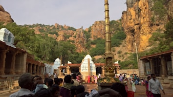 A group of people is gathered in the courtyard of a temple set against a rocky landscape. The architecture features ornate stone structures with white-topped towers. A tall, golden pillar stands prominently in the center, adorned with garlands. Lush greenery complements the rocky cliffs in the background.