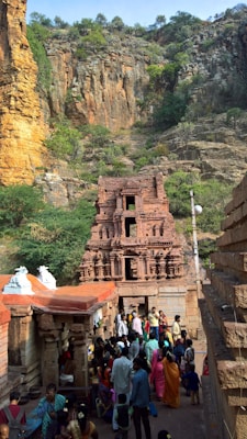 A group of people gathers near a historical stone temple situated against a rugged cliff with sparse greenery. The temple is adorned with intricate carvings and has a reddish-brown hue that complements the rock face. Brightly colored traditional clothing worn by the people adds vibrancy to the scene.