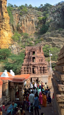 A group of people gathers near a historical stone temple situated against a rugged cliff with sparse greenery. The temple is adorned with intricate carvings and has a reddish-brown hue that complements the rock face. Brightly colored traditional clothing worn by the people adds vibrancy to the scene.