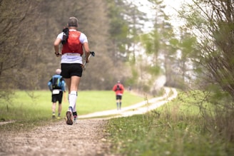 Several people are running along a winding gravel path in a forested area. The foreground features a runner in a red vest with white and black attire. The scene is lush with greenery, and the path stretches into the distance, surrounded by trees and grass.