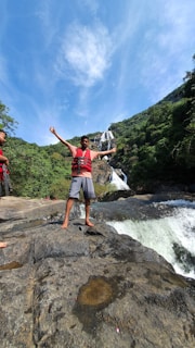 A person wearing a red life jacket stands on rocks near a waterfall, with arms outstretched. The surrounding area is lush with greenery, and the sky is clear with a few clouds.