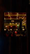 A warmly lit restaurant interior featuring elegant decor with columns, red lamps, and ceiling lights decorated with strings of small glowing bulbs. The ambiance is cozy and inviting, with tables set for dining and some visible patrons.