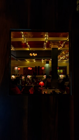 A warmly lit restaurant interior featuring elegant decor with columns, red lamps, and ceiling lights decorated with strings of small glowing bulbs. The ambiance is cozy and inviting, with tables set for dining and some visible patrons.