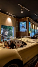 A classic, vintage car is parked indoors, featuring a polished white exterior and an open-top design. The car's dashboard is equipped with several dials and a wooden steering wheel. The interior environment is dimly lit with spotlights, while a large window offers a view of trees outside. There is also a framed picture of another car on the wall.